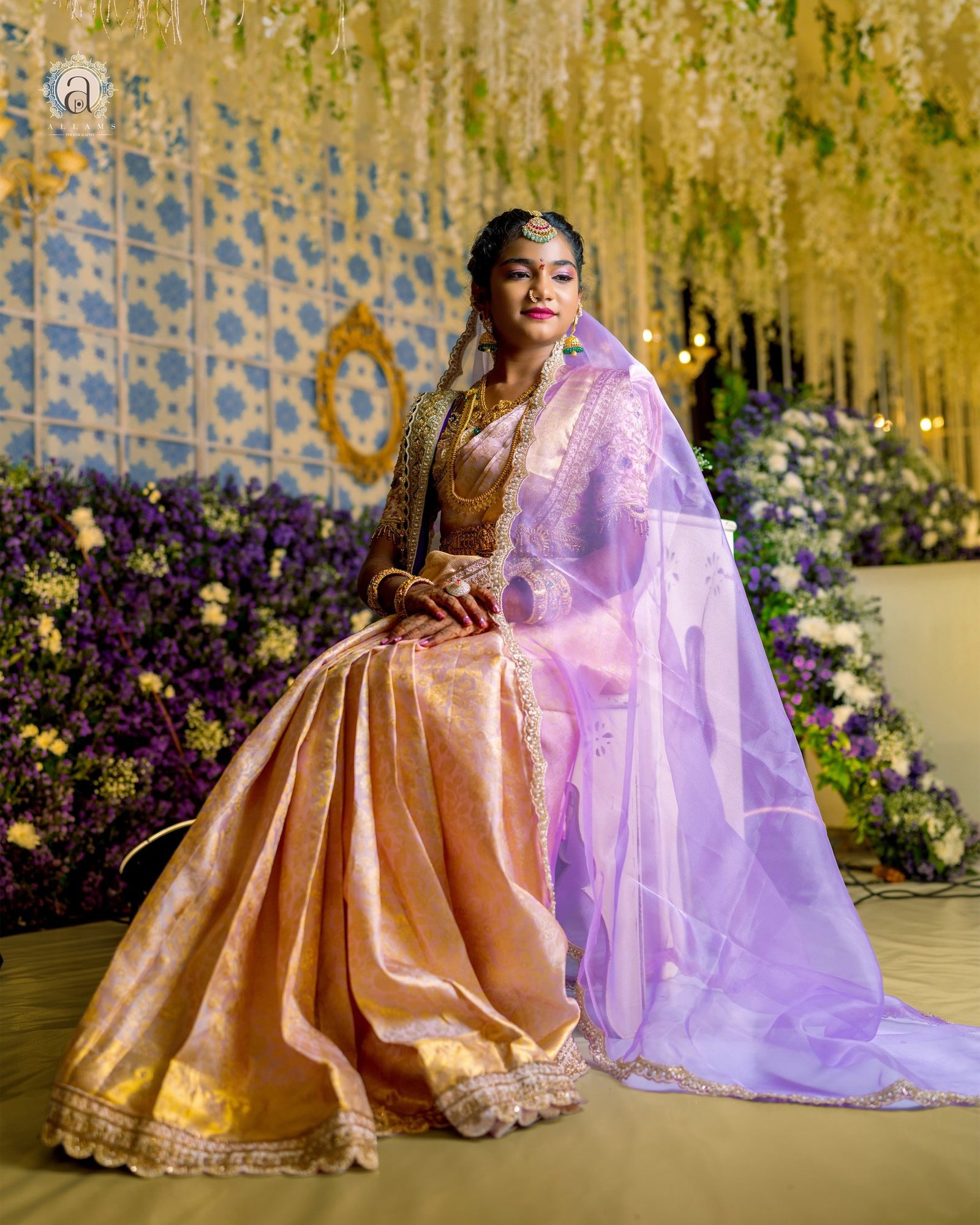A woman in a peach silk saree and lavender veil, seated before a flower wall.