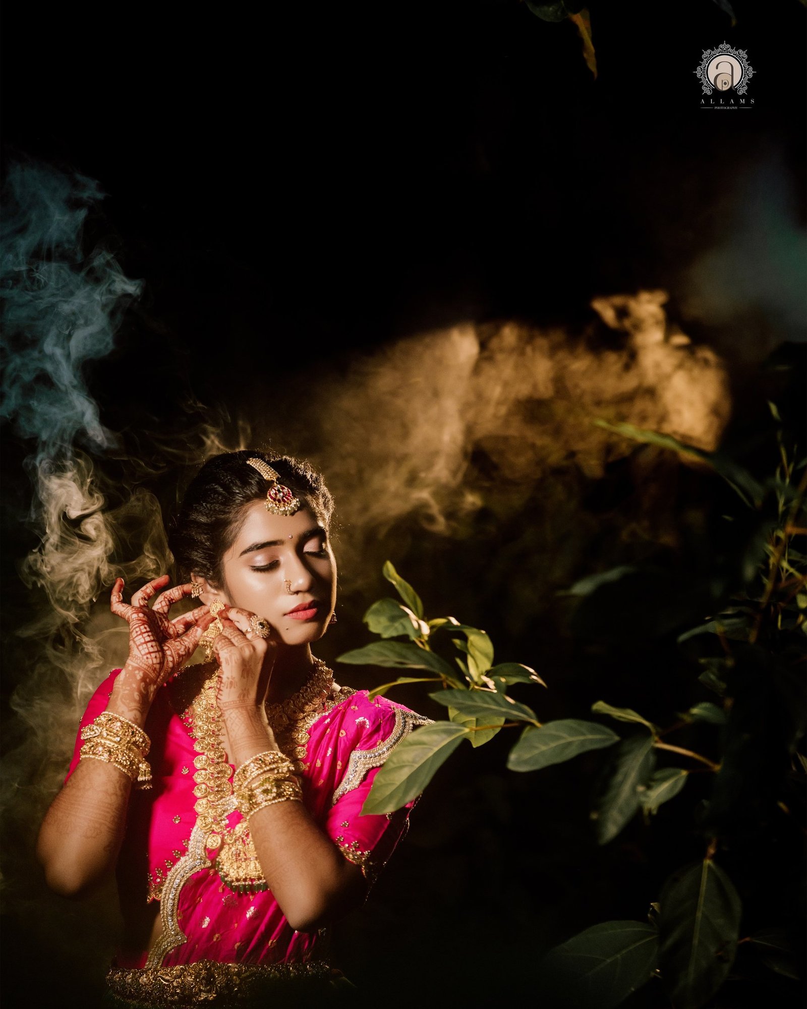 A woman in a bright pink saree and gold jewellery, hands raised near her face.
